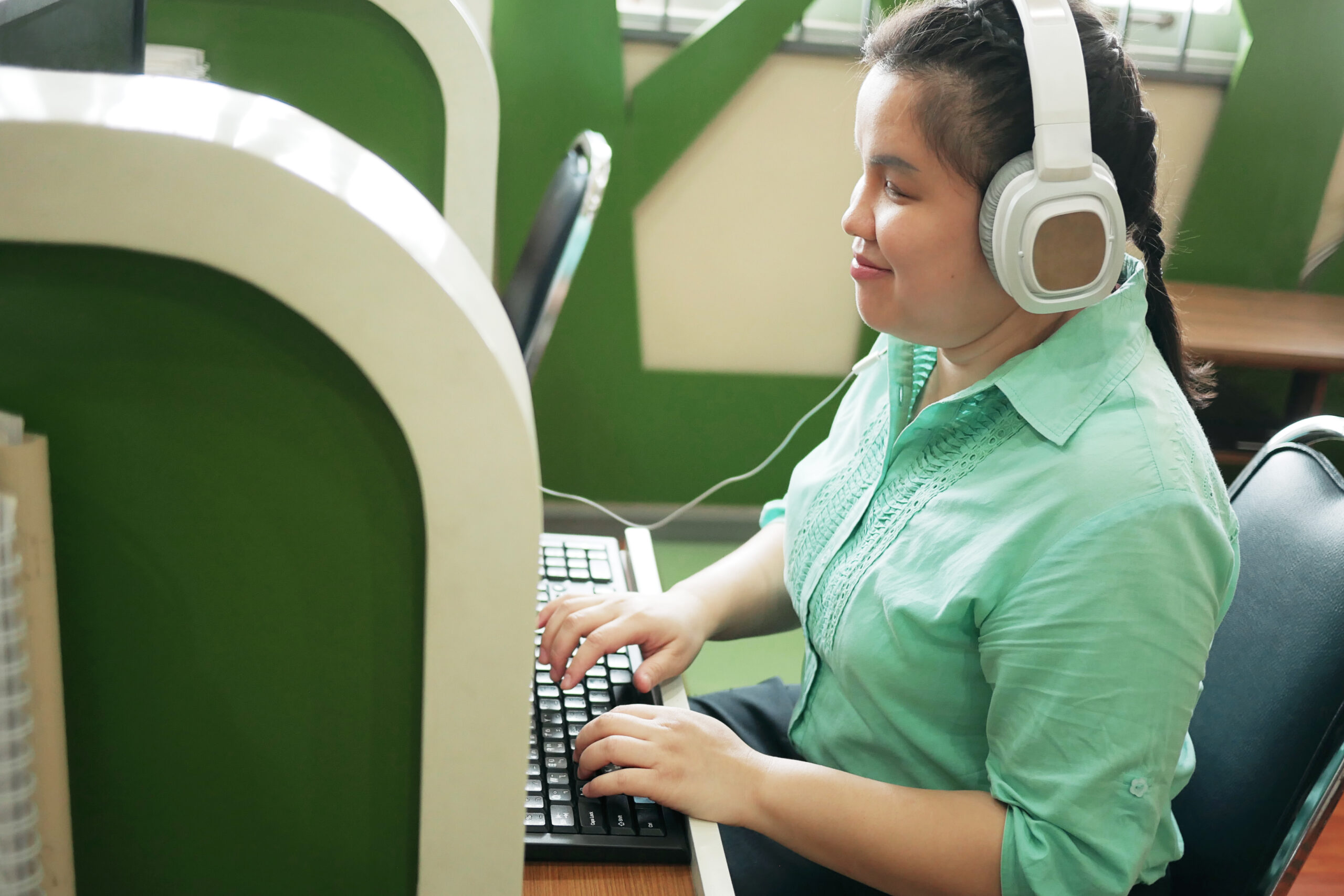 Blind woman sitting at computer cubicle, using computer, with headphones on.